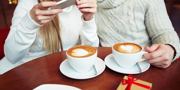 Young dates in sweaters photographing cappuccino while sitting in cafe